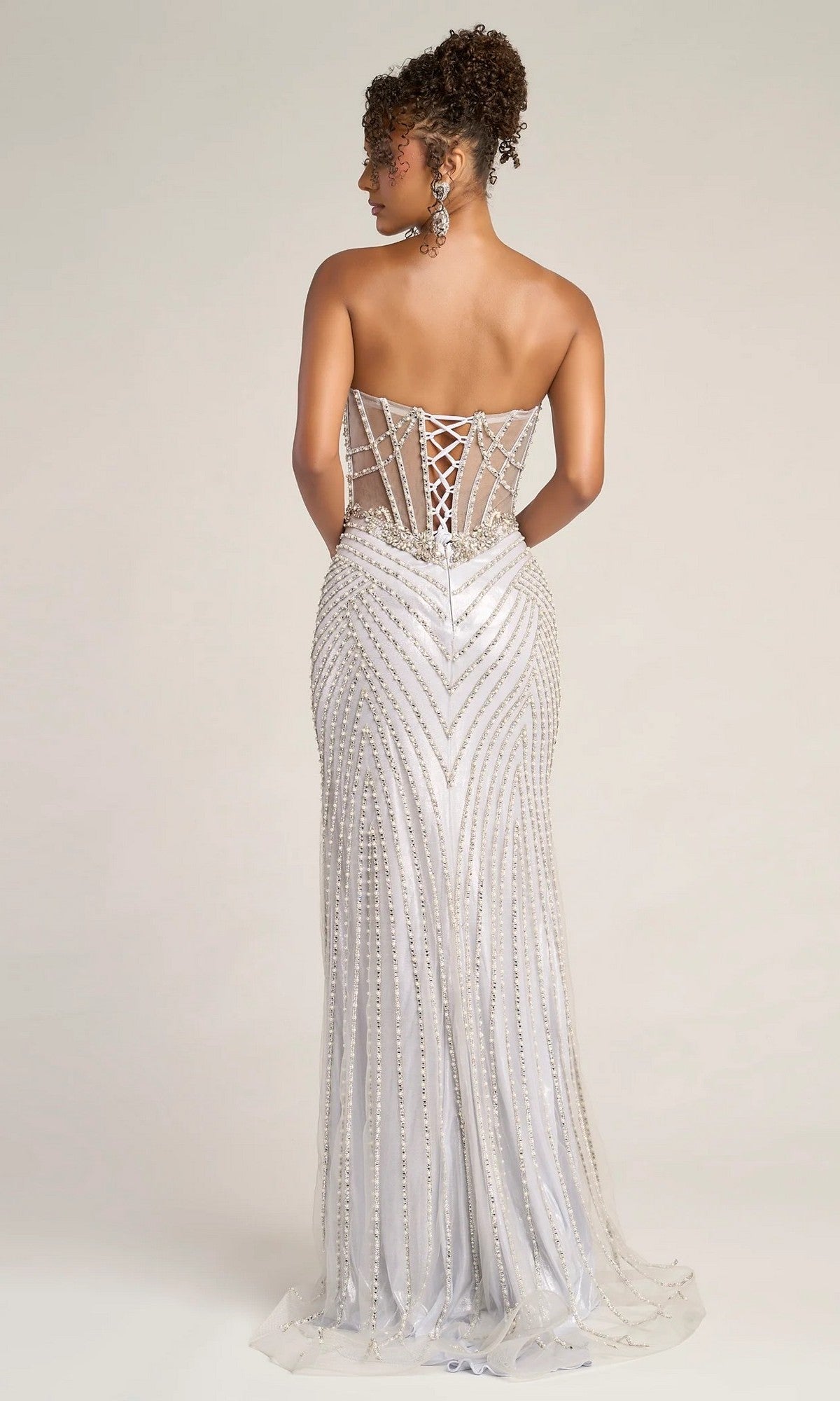 A woman models the Portia and Scarlett XO057, a strapless silver long formal dress with intricate beadwork and a lace-up corset back, standing against a plain light background.